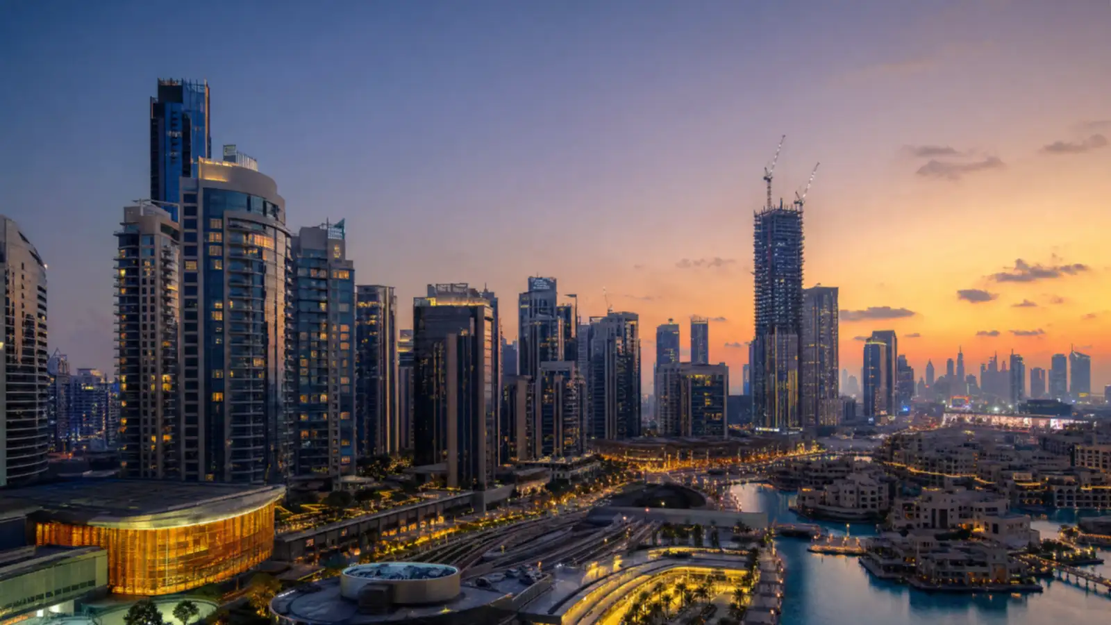 Dusk skyline combining a completed tower cluster with a crane-topped tower under construction in the distance